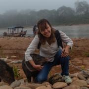 A woman with brown hair and bangs hugs one brown animal while another is to her right. The foreground has rocks and sandy dirt, and in the background is a boat, a river, trees and fog.